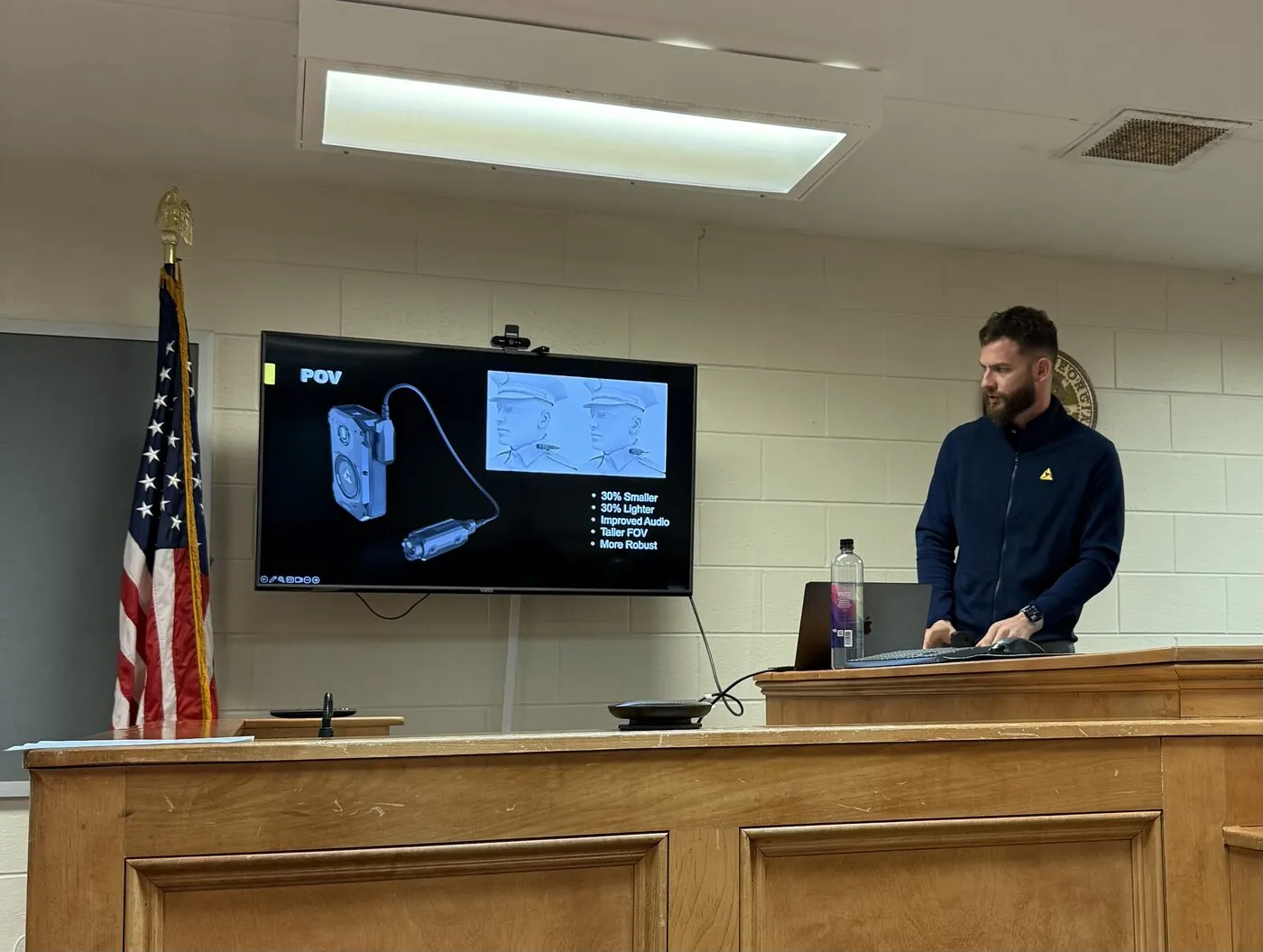 Shamir demoing an Axon body camera in a wood-paneled chamber, American flag in the background.